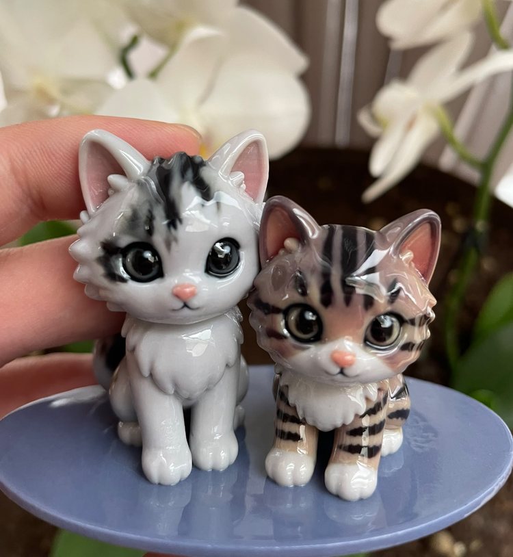 Two kitten figurines, one grey tabby and one striped, standing side by side on a lilac base